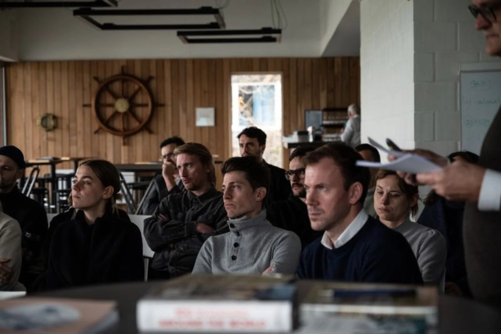 Deelnemers luisteren naar een presentatie in een zaal met houten wand en scheepsroer